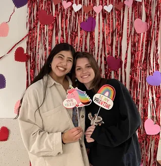 Two members of Medleys A Cappella posing with Valentine's Day cutouts against a backdrop of red streamers and various hearts